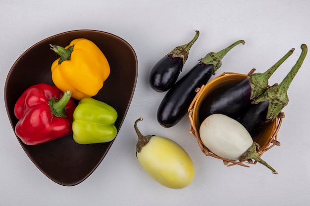 Vista superior berenjenas blancas y negras en una canasta con pimientos de colores en un recipiente sobre un fondo blanco.