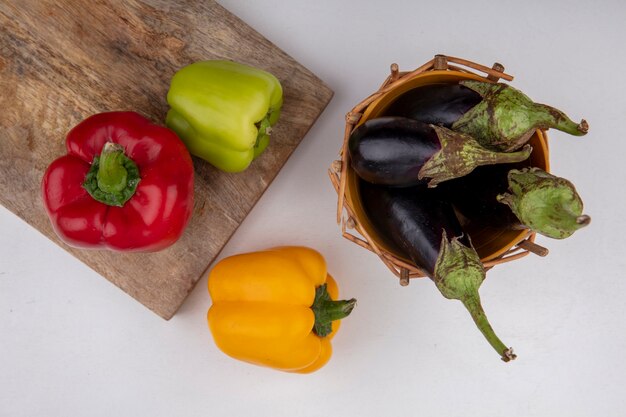 Vista superior berenjenas negras en una canasta con pimientos de colores sobre una tabla de cortar