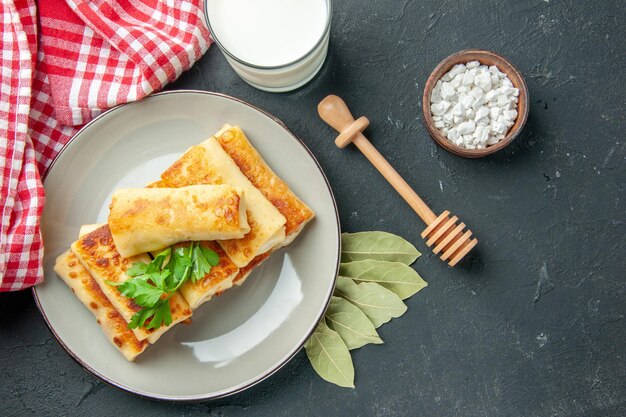 Vista superior de blini rellenos de carne en un plato redondo, sal marina en un tazón pequeño, hojas de laurel, palito de miel, vaso de suero de leche en la mesa