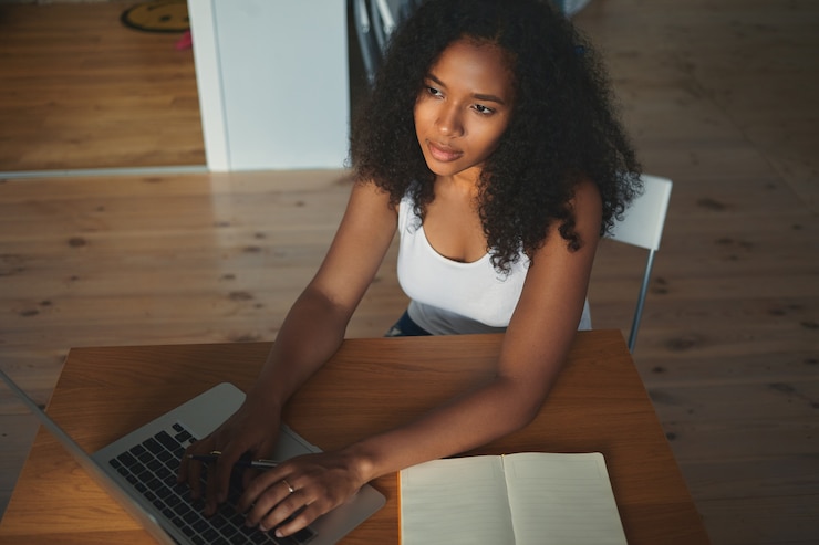 Foto gratuito vista superior de una bloguera de raza mixta joven pensativa y pensativa con cabello voluminoso que trabaja en un nuevo artículo para su blog, sentada en el escritorio frente a una computadora portátil abierta y escribiendo con el teclado