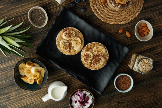 Vista superior de bocadillos de pan tostado con mantequilla de maní y plátano en un plato con queso cottage, leche, sésamo, avena, almendras, hojas de piña sobre fondo de madera