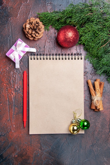 Vista superior bolígrafo rojo un cuaderno ramas de pino árbol de navidad juguetes de bolas palitos de canela en la superficie de color rojo oscuro foto de navidad