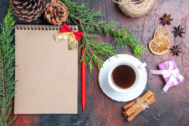 Vista superior bolígrafo rojo un cuaderno ramas de pino árbol de navidad juguetes y regalos de bolas una taza de té platillo blanco palitos de canela anís sobre superficie roja oscura