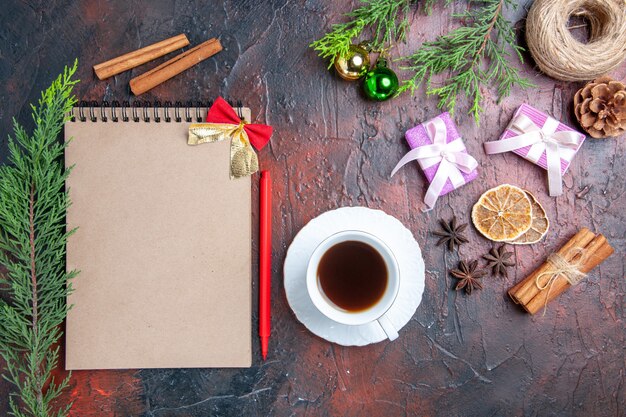 Vista superior bolígrafo rojo un cuaderno ramas de pino árbol de navidad juguetes y regalos una taza de té platillo blanco anís canela sobre superficie roja oscura