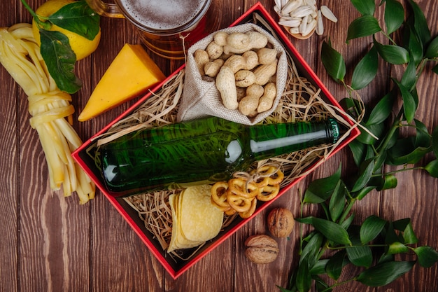 Vista superior de la botella de cerveza con maní en un saco de papas fritas y mini pretzels en paja en una caja roja y una jarra de cerveza con queso y limón en rústico