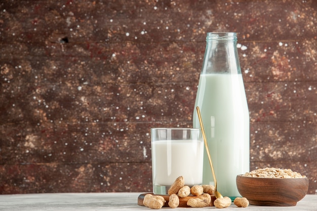 Vista superior de la botella de vidrio y la taza llena de leche en la bandeja de madera y frutos secos cuchara avena en una olla marrón en el lado izquierdo de la mesa blanca sobre fondo marrón