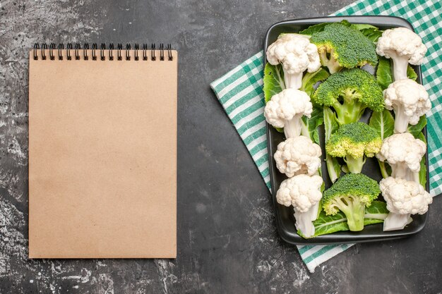 Vista superior de brócoli y coliflor en un plato rectangular negro sobre una servilleta a cuadros verde y blanca un cuaderno sobre fondo oscuro foto de comida