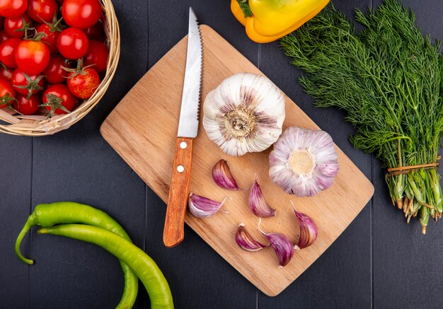 Vista superior de bulbo de ajo y dientes con cuchillo en la tabla de cortar y una cesta de tomate en superficie negra