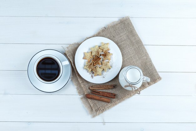 Vista superior de café en taza con galletas, canela, leche en madera y un pedazo de fondo de saco.