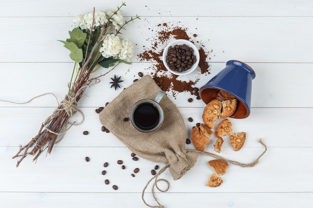 Vista superior de café en taza con granos de café, galletas, flores sobre fondo de madera y saco.