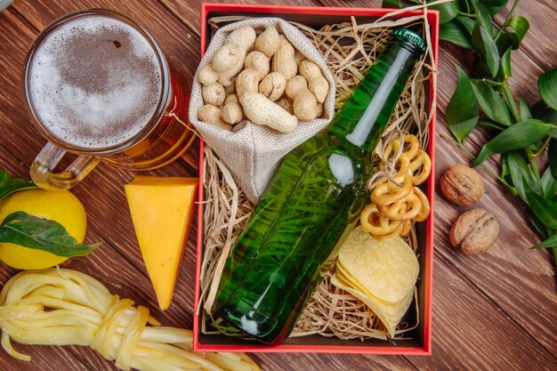 Vista superior de una caja con una botella de cerveza, maní, papas fritas, mini pretzels y paja rústica con una taza de cerveza, queso y limón.