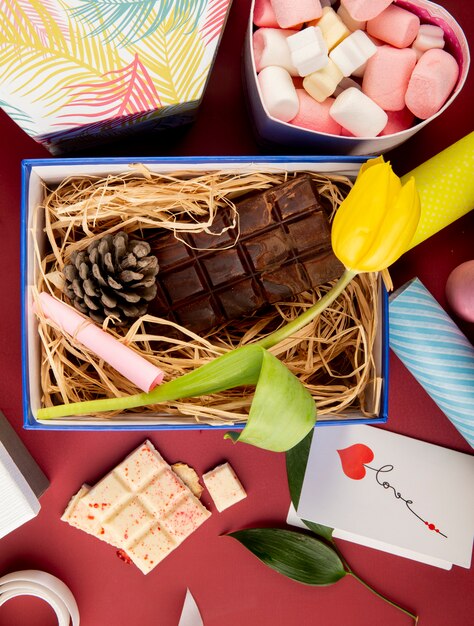 Vista superior de una caja de regalo abierta con flor de tulipán de color amarillo, barra de chocolate oscuro, cono y paja y una caja en forma de corazón llena de malvaviscos en la mesa de color rojo oscuro