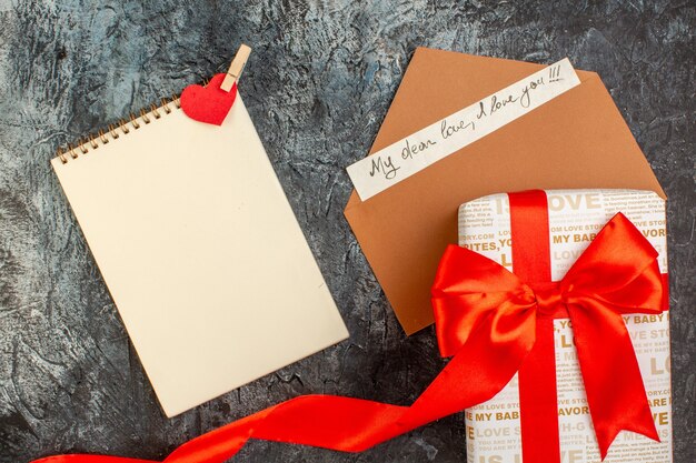 Foto gratuita vista superior de la caja de regalo bellamente empaquetada atada con cinta roja en un sobre con cuaderno de espiral de carta de amor sobre fondo oscuro helado