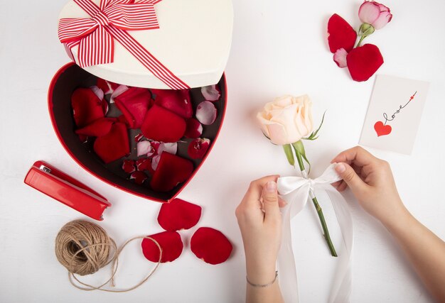 Vista superior de la caja de regalo en forma de corazón llena de pétalos de rosa roja grapadora de color rojo cuerda y manos femeninas atando una rosa blanca con una cinta sobre fondo blanco.