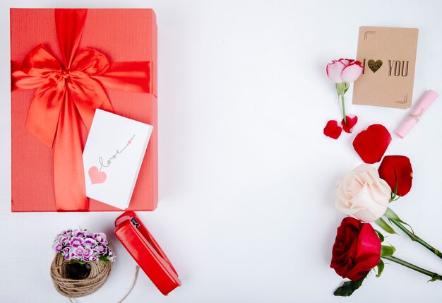 Vista superior de la caja de regalo roja con un lazo y rosas de color rojo y blanco con grapadora y pequeña postal sobre fondo blanco con espacio de copia