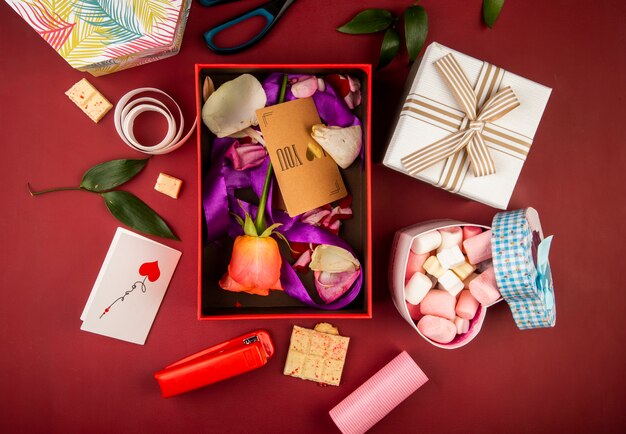 Vista superior de una caja de regalo roja con tarjeta de papel marrón y flor color de rosa coral y pétalos con cinta morada y caja en forma de corazón llena de malvavisco en la mesa de color rojo oscuro