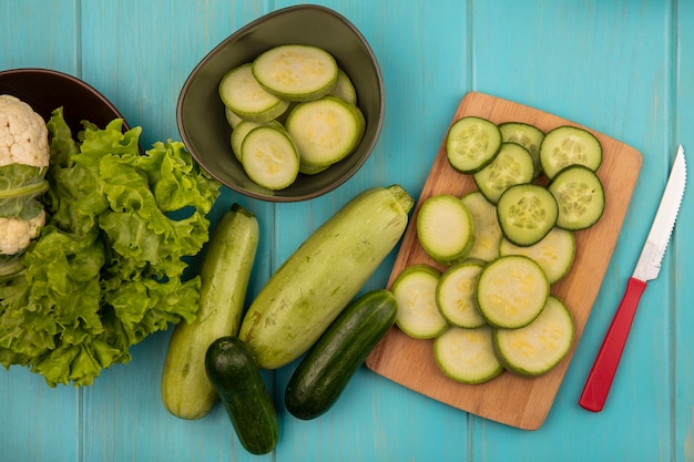 Vista superior de calabacines y pepinos picados en una tabla de cocina de madera con un cuchillo con coliflor y lechuga en un recipiente con pepinos enteros y calabacines aislados en una superficie de madera azul