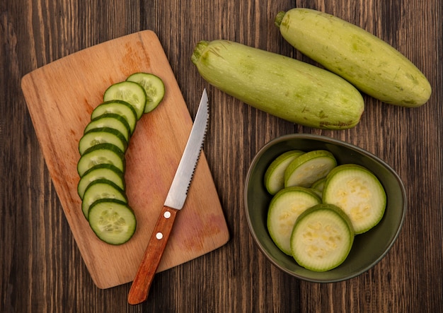 Vista superior de calabacines picados en un recipiente con pepinos picados sobre una tabla de cocina de madera con cuchillo con calabacines aislado en una pared de madera