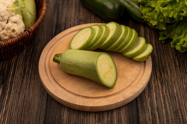 Vista superior de calabacines picados en una tabla de cocina de madera con verduras como calabacines y coliflor en un balde con lechuga y pepinos aislado en una pared de madera