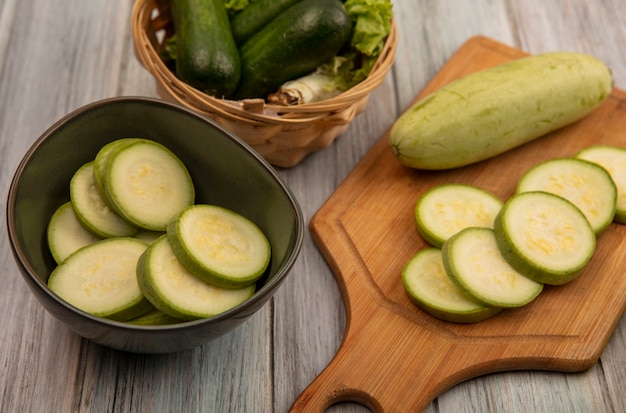 Vista superior de calabacines de piel verde sobre una tabla de cocina de madera con calabacines picados en un recipiente con pepinos y lechuga en un balde en una pared de madera gris