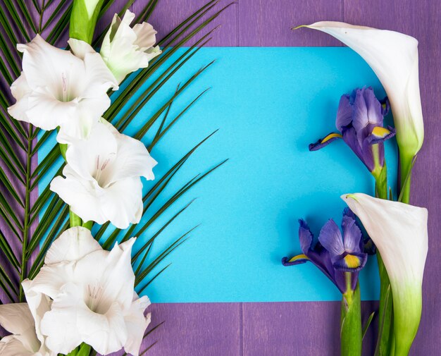 Vista superior de calas y gladiolos de color blanco con flor de iris morado oscuro y hoja de palma con una hoja de papel azul sobre fondo morado