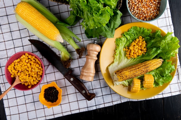 Vista superior de callos enteros y cortados y semillas de maíz con lechuga en un plato y espinacas cuchillo pimienta negra sobre tela escocesa y superficie negra