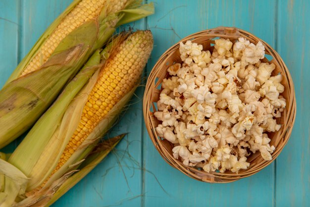 Vista superior de callos orgánicos y saludables con cabello con palomitas de maíz en un cubo sobre una mesa de madera azul