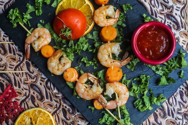 Vista superior de camarones a la parrilla en brochetas con zanahorias, tomate, hierbas y salsa en un soporte