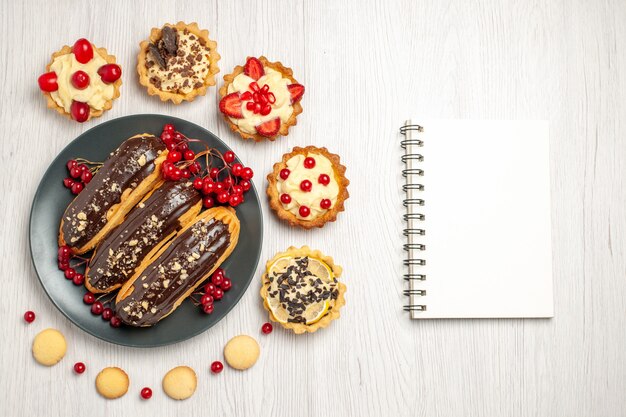 Vista superior canutillos de chocolate y pasas de Corinto en la placa gris rodeada de tartas y galletas y un cuaderno en la mesa de madera blanca con espacio de copia