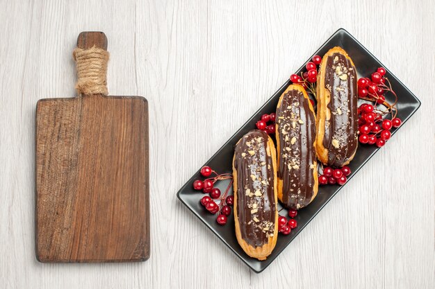 Vista superior de canutillos de chocolate y pasas de Corinto en el plato rectangular negro en el lado derecho y una tabla de cortar en el lado izquierdo del suelo de madera blanca