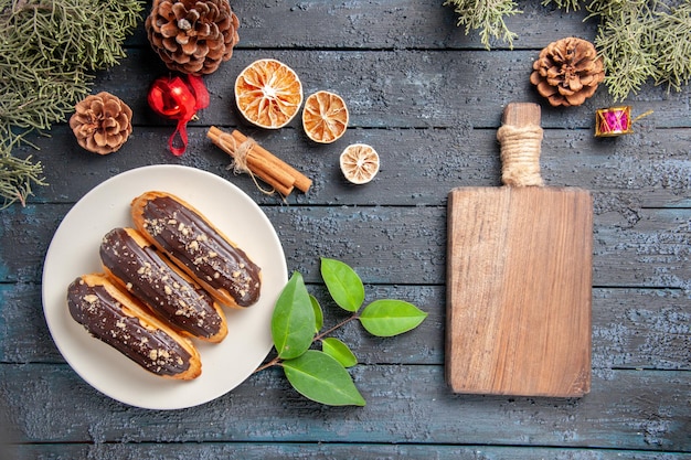 Vista superior canutillos de chocolate en una placa ovalada conos juguetes de navidad hojas de abeto naranjas secas de canela y tabla de cortar sobre suelo de madera oscura.