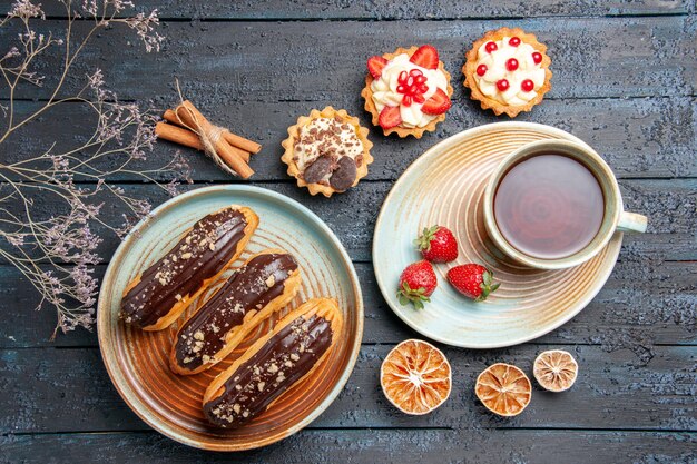 Vista superior canutillos de chocolate en placa ovalada una taza de té y fresas en tartas de platillo canela y limones secos en la mesa de madera oscura.