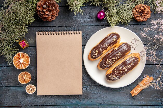 Vista superior canutillos de chocolate en plato ovalado blanco ramas de abeto y conos juguetes de navidad rama de flor seca naranjas secas canela y un cuaderno en la mesa de madera oscura