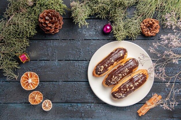 Vista superior canutillos de chocolate en plato ovalado blanco ramas de abeto y conos juguetes navideños naranjas secas canela en mesa de madera oscura con espacio libre