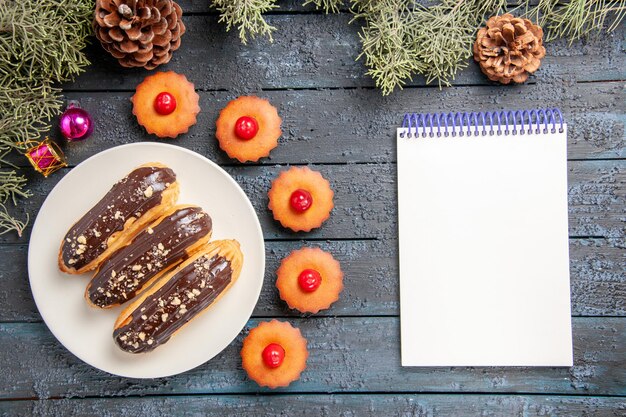 Vista superior canutillos de chocolate en plato ovalado blanco ramas de abeto juguetes de navidad cupcakes de cereza y un cuaderno en la mesa de madera oscura