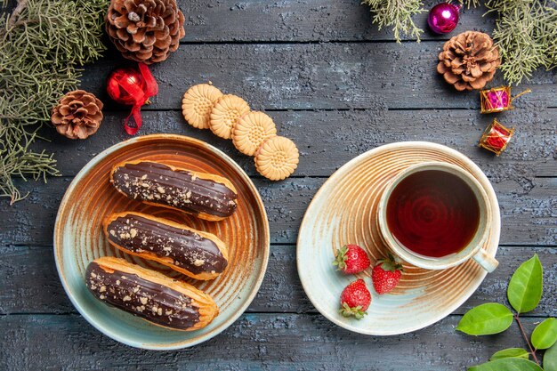 Vista superior canutillos de chocolate en un plato ovalado una taza de té y fresas en un platillo piñas juguetes navideños hojas de abeto galletas sobre suelo de madera oscura