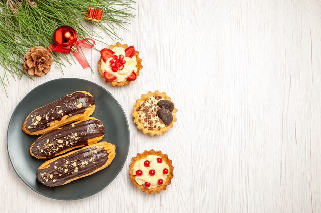 Vista superior de canutillos de chocolate en las tartas de placa gris y hojas de pino con juguetes navideños en el lado izquierdo del suelo de madera blanca