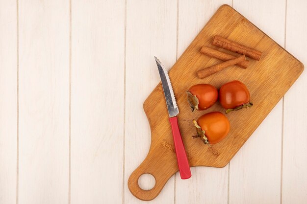 Vista superior de caquis suaves sobre una tabla de cocina de madera con ramas de canela con un cuchillo sobre una superficie de madera blanca con espacio de copia