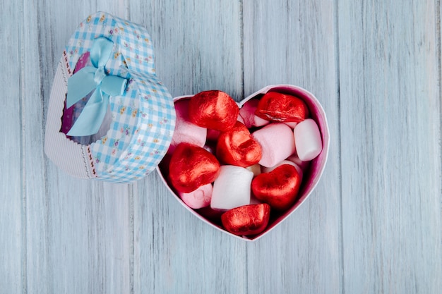 Vista superior de caramelos de chocolate en forma de corazón envueltos en papel rojo con malvavisco rosa en una caja de regalo en forma de corazón sobre una mesa de madera gris