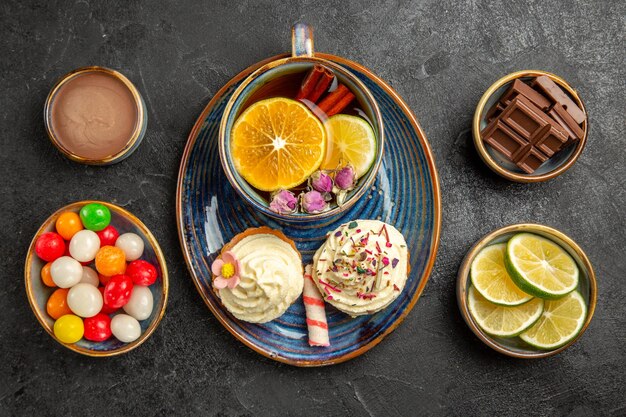 Vista superior de caramelos en la mesa cuencos de limas y dulces junto al platillo de dos cupcakes y la taza de té con palitos de canela y limón sobre la mesa