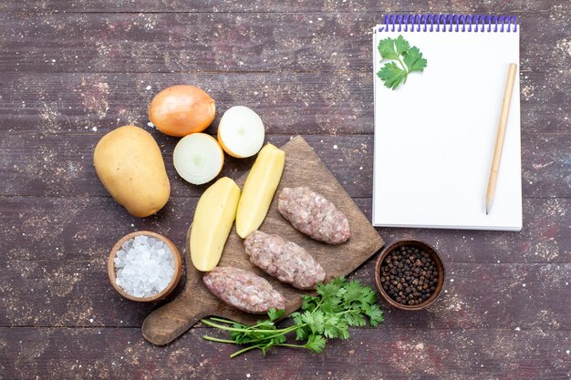 Vista superior de carne cruda con patatas crudas, sal, cebolla, bloc de notas y verduras en el escritorio marrón, carne, plato de patatas, comida, cena