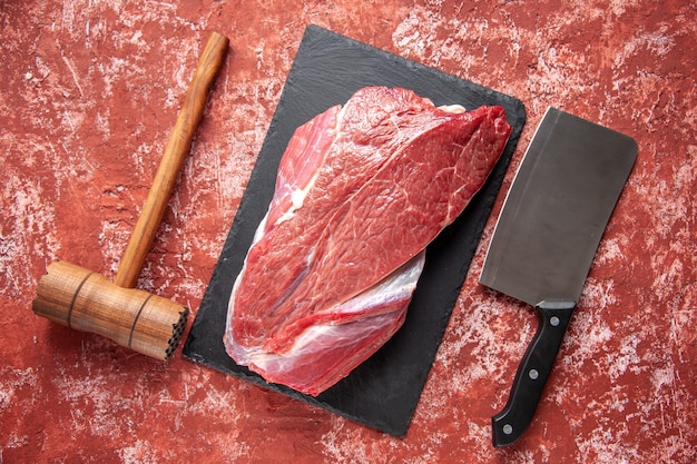 Vista superior de carne fresca cruda roja en tablero negro martillo de madera marrón y hacha sobre fondo rojo pastel