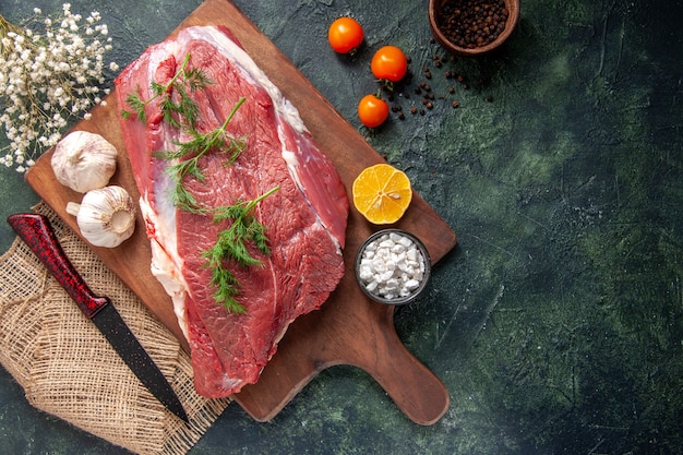 Vista superior de carnes rojas crudas frescas, ajos verdes, sal de limón en un cuchillo de tabla de cortar de madera marrón en una toalla de color nude, tomates, pimienta sobre fondo de color oscuro