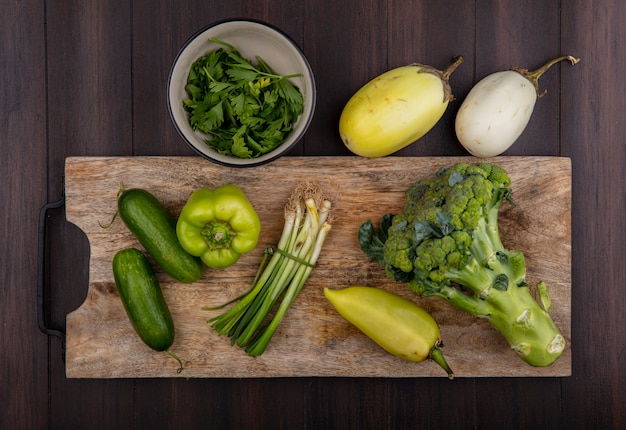 Vista superior de cebollas verdes con pepinos y pimientos verdes brócoli sobre una tabla de cortar con perejil en un recipiente sobre un fondo de madera