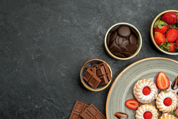 Vista superior de cerca apetitosas galletas dulces con fresa y tazones de fresa y chocolate en el lado derecho de la mesa oscura