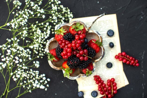 Una vista superior de cerca frutas frescas bayas coloridas alrededor de papel y flores blancas en la superficie oscura