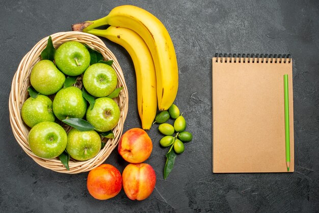 Vista superior de cerca frutas plátanos limas manzanas en la canasta nectarinas junto al lápiz de cuaderno
