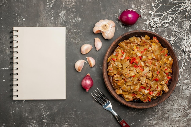 Vista superior de cerca judías verdes con tomates plato de madera de judías verdes y tomates junto a la cebolla, ajo, cuaderno blanco y tenedor sobre la mesa