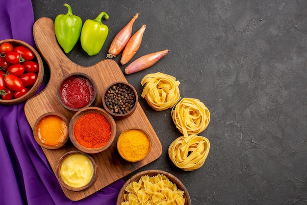 Vista superior de cerca pasta y tomates especias y salsas en tazones en la tabla de cortar bola pimiento cebolla tazón de tomates y pasta en el mantel morado sobre la mesa