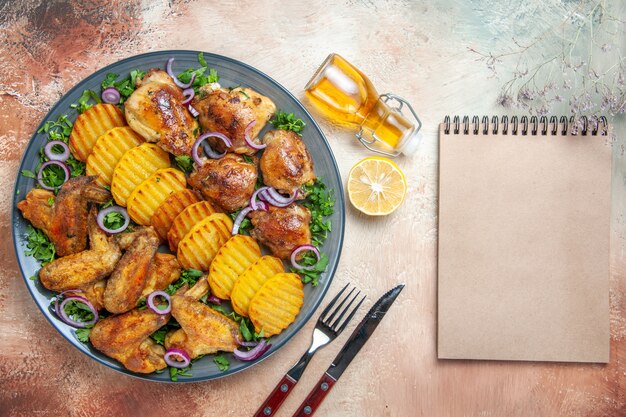 Vista superior de cerca pollo alitas de pollo patatas hierbas aceite de cebolla limón tenedor cuchillo crema cuaderno